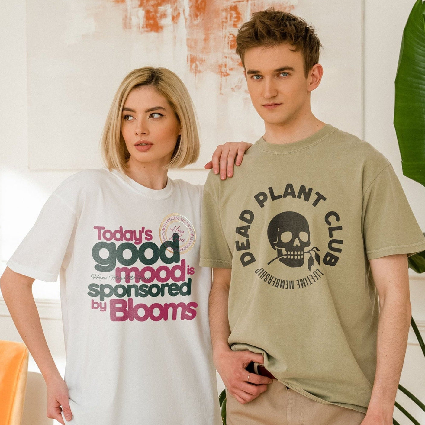 Two models wearing graphic T-shirts from the Dusty Aloe shop. The woman wears an oversized white shirt with the phrase “Today’s good mood is sponsored by Blooms” in bold colorful letters. The man wears a light olive-green shirt with a skull graphic and the words “Dead Plant Club.” They are standing indoors beside a tall green plant.